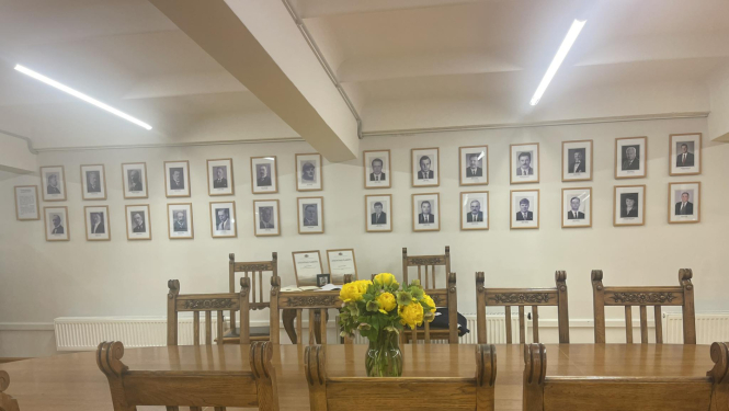 Kabinets (telpa) ar lielu galdu un krēsliem. Uz galda vāzē dzeltenas puķes, pie sienas Latvijas Republikas zemkopības ministru foto galerija.