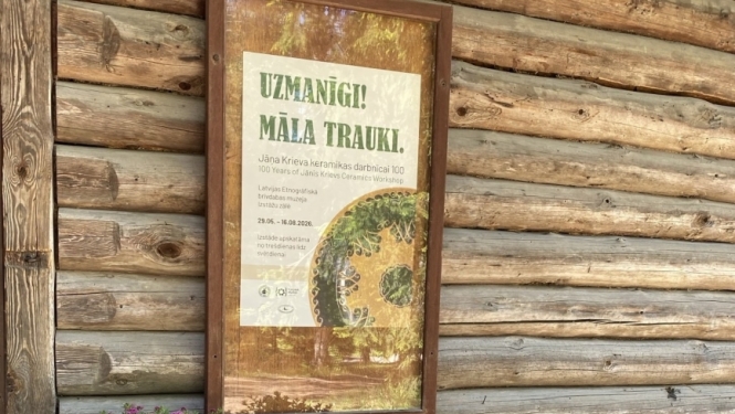 Pie guļbūves sienas pielikta afiša par māla trauku izstādi. Pie sienas ziedu krūms rozā toņos