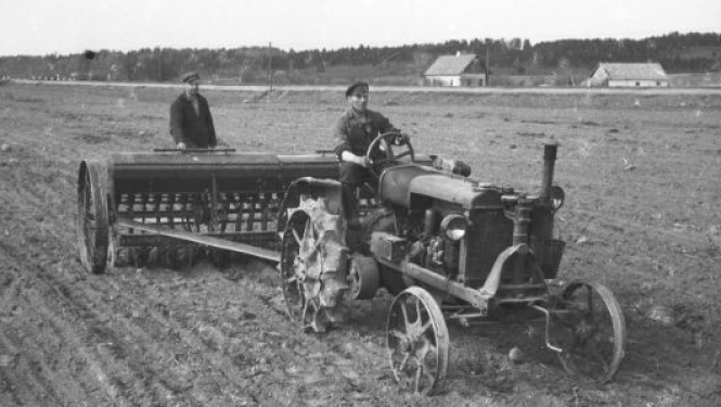 Muzeja izstādes informatīvais stends. Vidū bilde - senlaicīgs traktors velk sējmašīnu, kurā sēklu regulē cilvēks. Fons zaļos toņos, zāle tuvplānā.