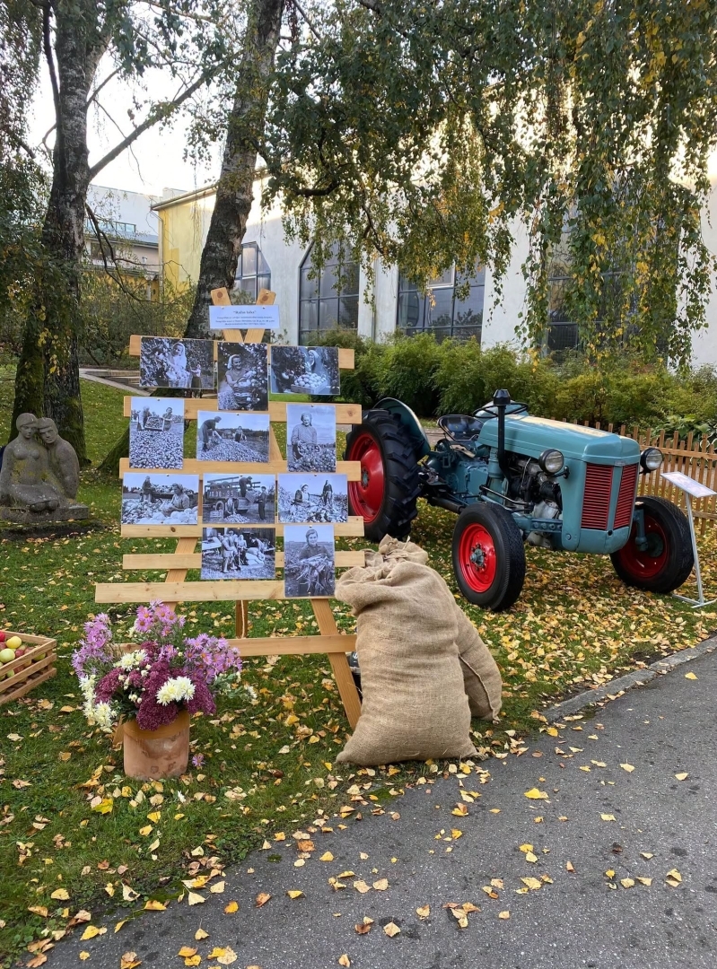 Lapkritis zem bērziem, kurā nolikts senlaicīgs traktors, brūni maisi un senlaicīgas fotogrāfijas