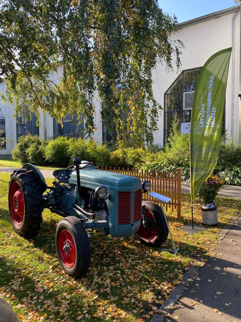 Riteņtraktors Ferguson TEF20 novietots uz nokritušām bērza lapām. Fonā liela gaiša ēka un muzeja karogs