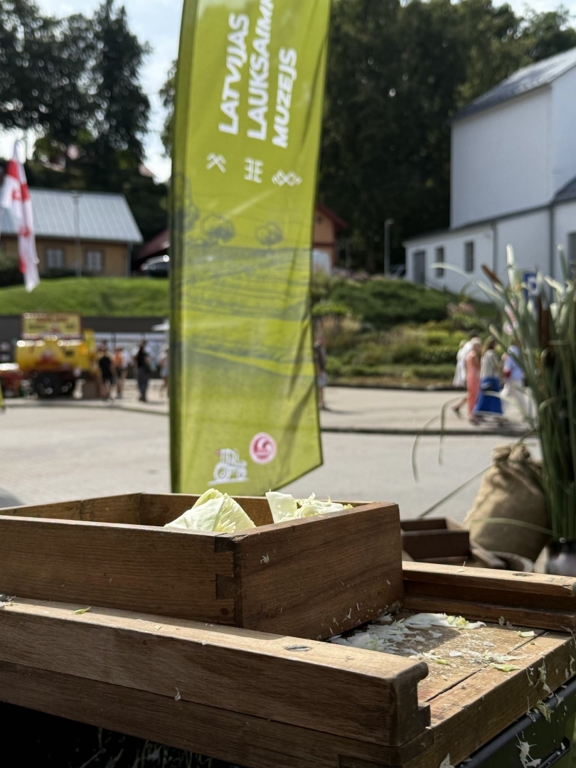 Priekšplānā kāpostu ēvele, aiz kuras muzeja karogs un raibi cilvēki Dižmāras gadatirgū Talsos