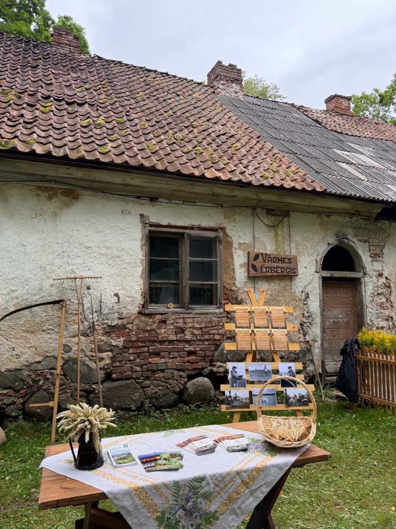 Tematiska ainava - fonā mūra ēka, priekšā ar raibu galdautu klāts galds. Pie mājas piestutēti senlaicīgi darbarīki.