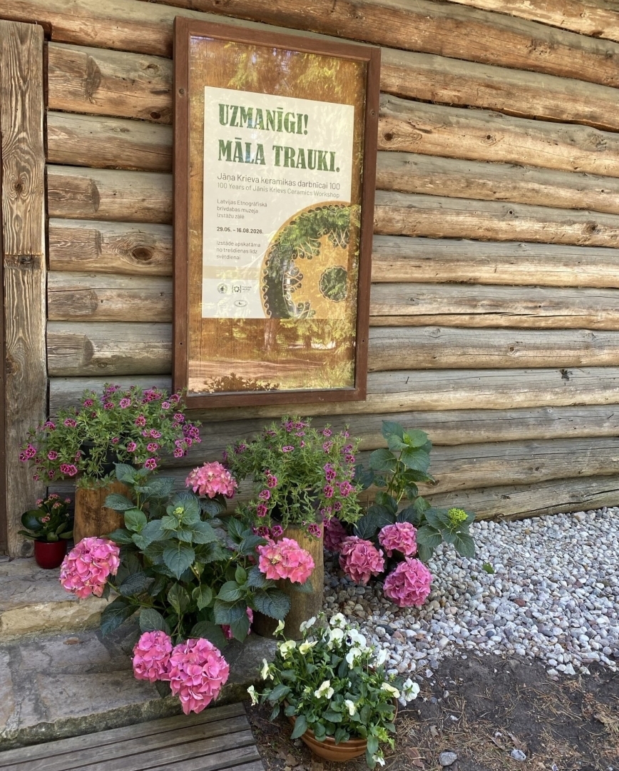 Pie guļbūves sienas pielikta afiša par māla trauku izstādi. Pie sienas ziedu krūms rozā toņos