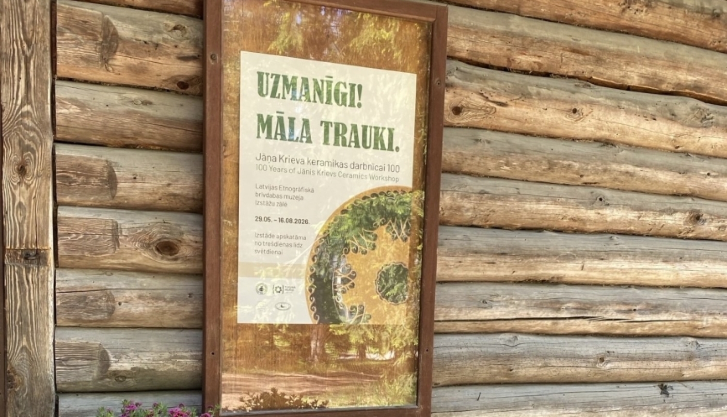 Pie guļbūves sienas pielikta afiša par māla trauku izstādi. Pie sienas ziedu krūms rozā toņos