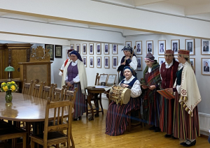 Muzeja telpās pie ekspozīcijas redzamas sievietes no folkloras kopas, muzicē