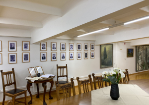 Ekspozīcijas skats muzeja telpā, ar krēsliem, galdu un ziediem uz tā, portretu galeriju pie sienas