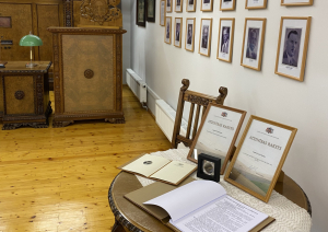 Ekspozīcijas skats muzeja telpā, ar senlaicīgām mēbelēm, mazu galdu un atzinības materiāliem uz tā, portretu galeriju pie sienas