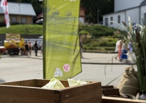 Priekšplānā kāpostu ēvele, aiz kuras muzeja karogs un raibi cilvēki Dižmāras gadatirgū Talsos