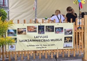 Latvijas Lauksaimniecības muzeja baneris uz gaišas koka sētas. Fonā divas meitenes un vēja ķērājs bites formā