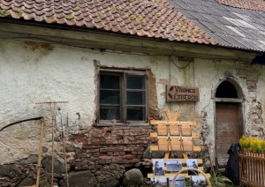 Tematiska ainava - fonā mūra ēka, priekšā ar raibu galdautu klāts galds. Pie mājas piestutēti senlaicīgi darbarīki.
