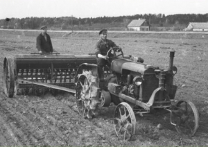 Muzeja izstādes informatīvais stends. Vidū bilde - senlaicīgs traktors velk sējmašīnu, kurā sēklu regulē cilvēks. Fons zaļos toņos, zāle tuvplānā.