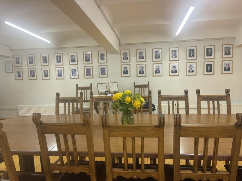 Kabinets (telpa) ar lielu galdu un krēsliem. Uz galda vāzē dzeltenas puķes, pie sienas Latvijas Republikas zemkopības ministru foto galerija.