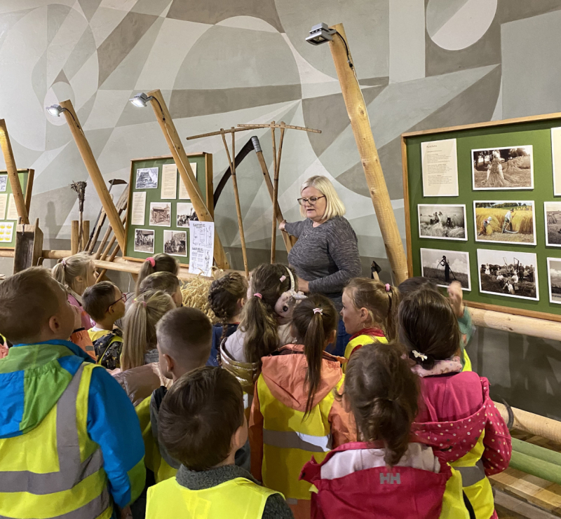 Sieviete gaišiem matiem bērnu bariņā muzeja telpās. Bērni saģērbti zaļās, atstarojošās vestēs