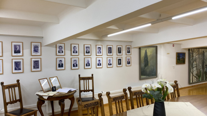 Ekspozīcijas skats muzeja telpā, ar krēsliem, galdu un ziediem uz tā, portretu galeriju pie sienas