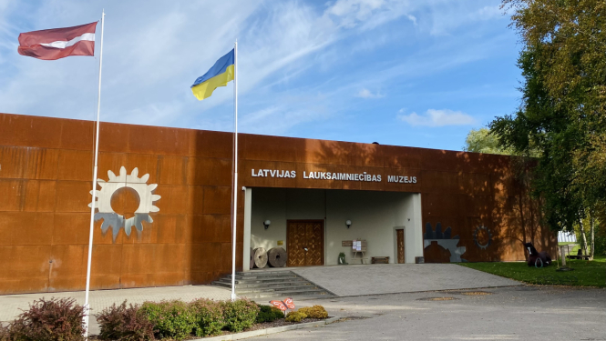 Latvijas Lauksaimniecības muzeja ēkas ieejas skats, ar diviem plīvojošiem karogiem un oranžu taureni pie karogu mastiem