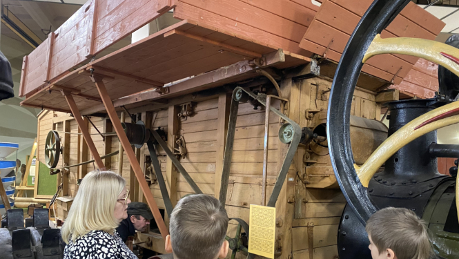 Divi skolnieki muzeja telpās klausās vienā pieaugušajā par senlaicīgu kuļmašīnu