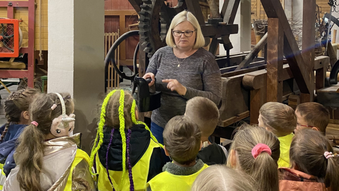 Sieviete gaišiem matiem bērnu bariņā muzeja telpās. Bērni saģērbti zaļās, atstarojošās vestēs