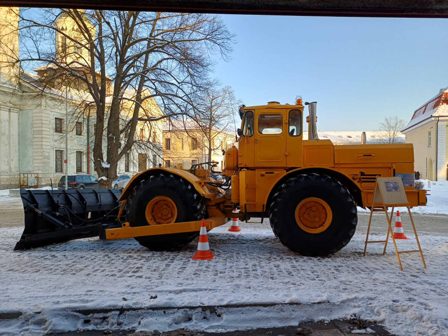 Liels dzeltens traktors ar lāpstu sniegotā pilsētvidē