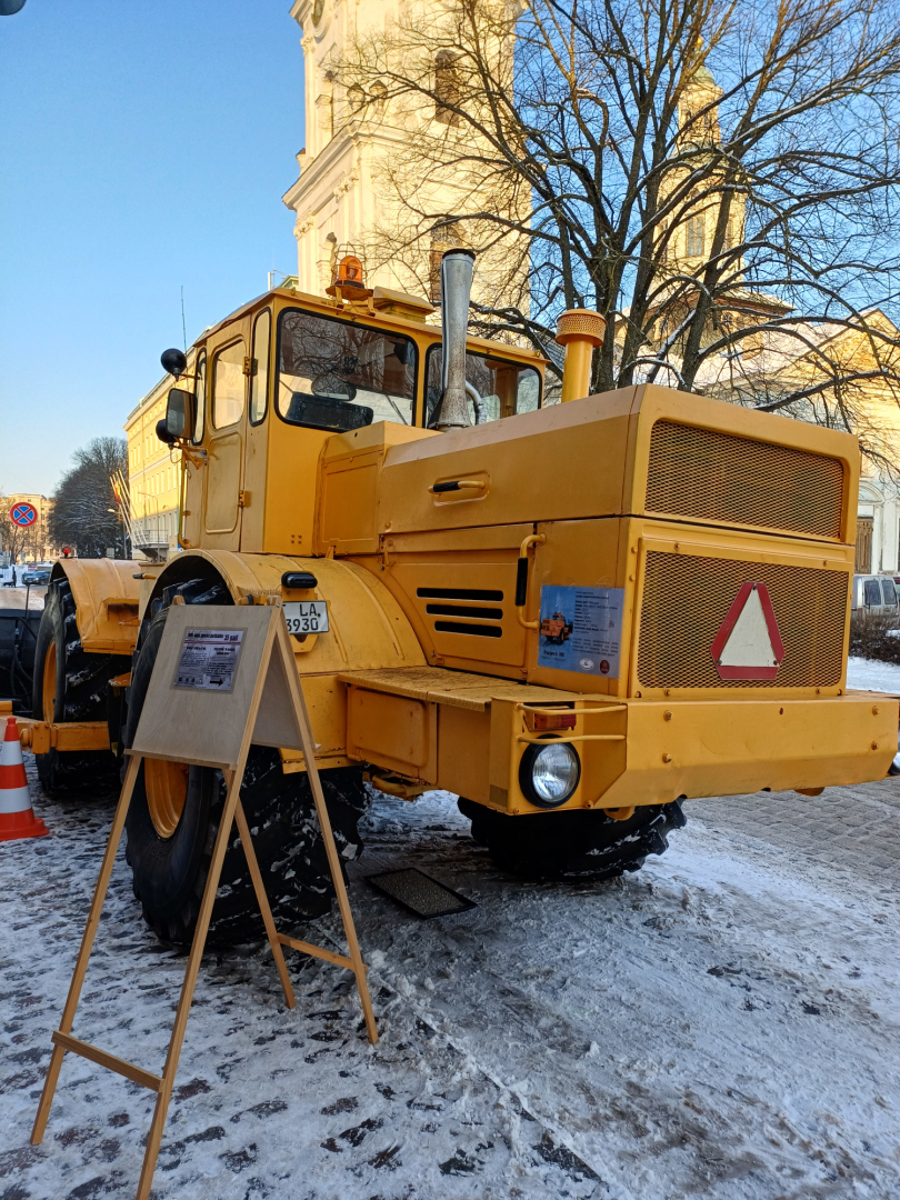 Liels dzeltens traktors ar lāpstu sniegotā pilsētvidē