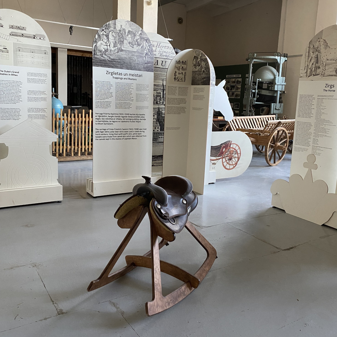 Muzeja telpās fonā vairāki, gareni informatīvi stendi par zirgu un zirgošanās tēmu, centrā apseglotas koka zirga šūpoles bez galvas
