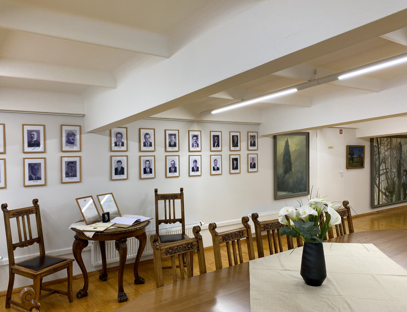 Ekspozīcijas skats muzeja telpā, ar krēsliem, galdu un ziediem uz tā, portretu galeriju pie sienas