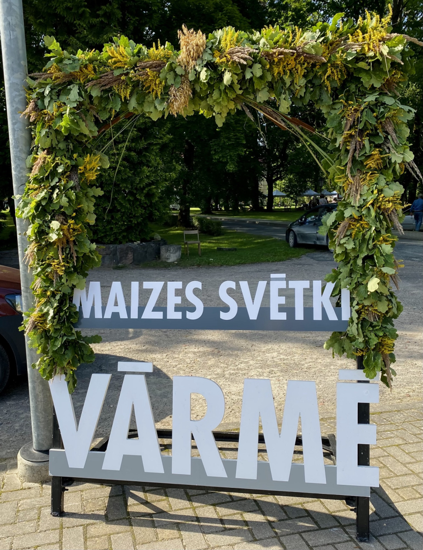 Dabas materiāliem izrotāts uzraksts maizes svētki Vārmē. Fonā bruģēts ieejas laukums parkā, pasākuma norises vietā