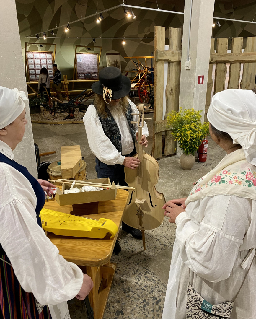 Divas sievietes baltās aubēs un viens kungs melnā cepurē demonstrē tautas mūzikas instrumentu izgatavošanas meistarklasi