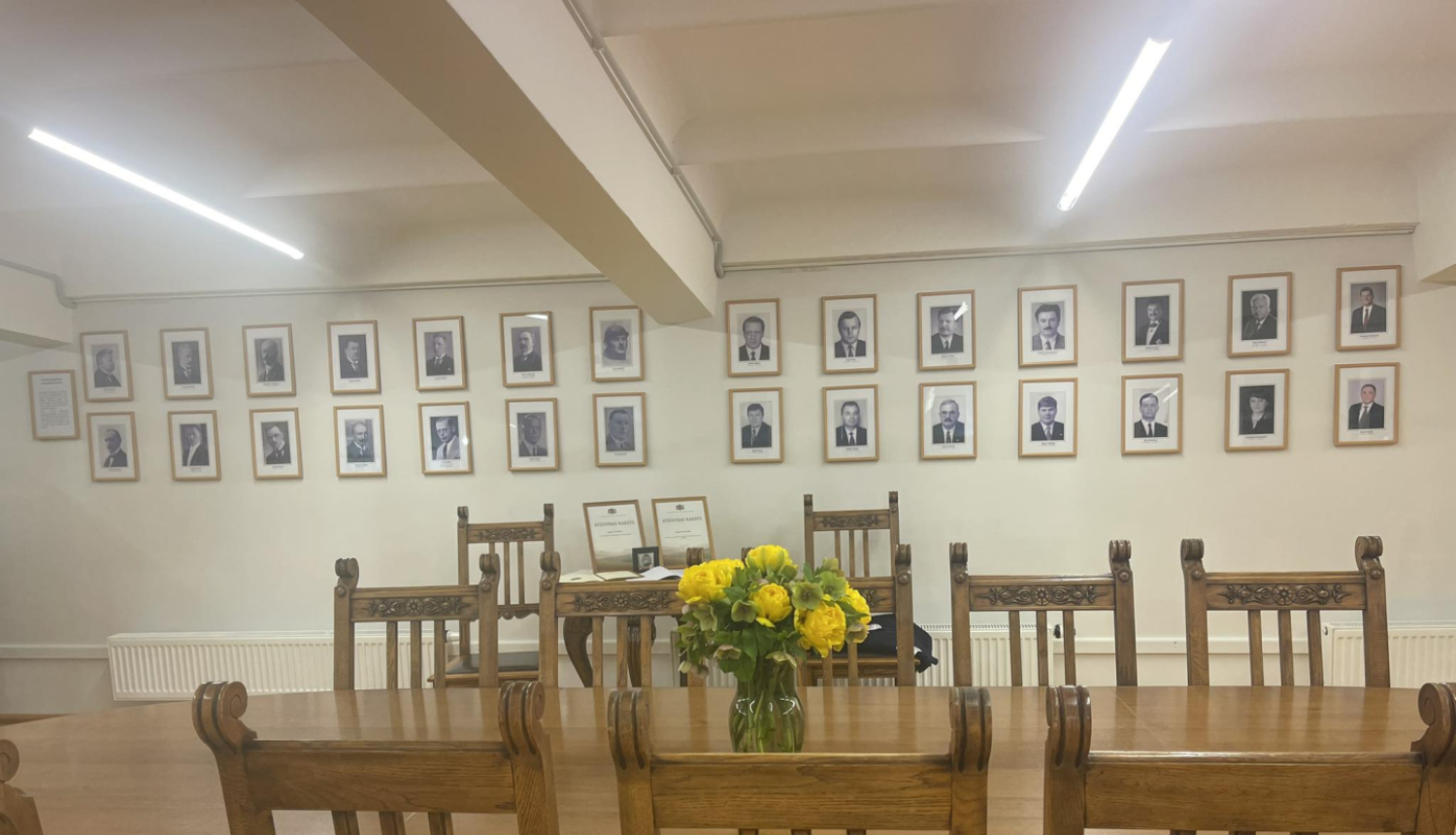 Kabinets (telpa) ar lielu galdu un krēsliem. Uz galda vāzē dzeltenas puķes, pie sienas Latvijas Republikas zemkopības ministru foto galerija.