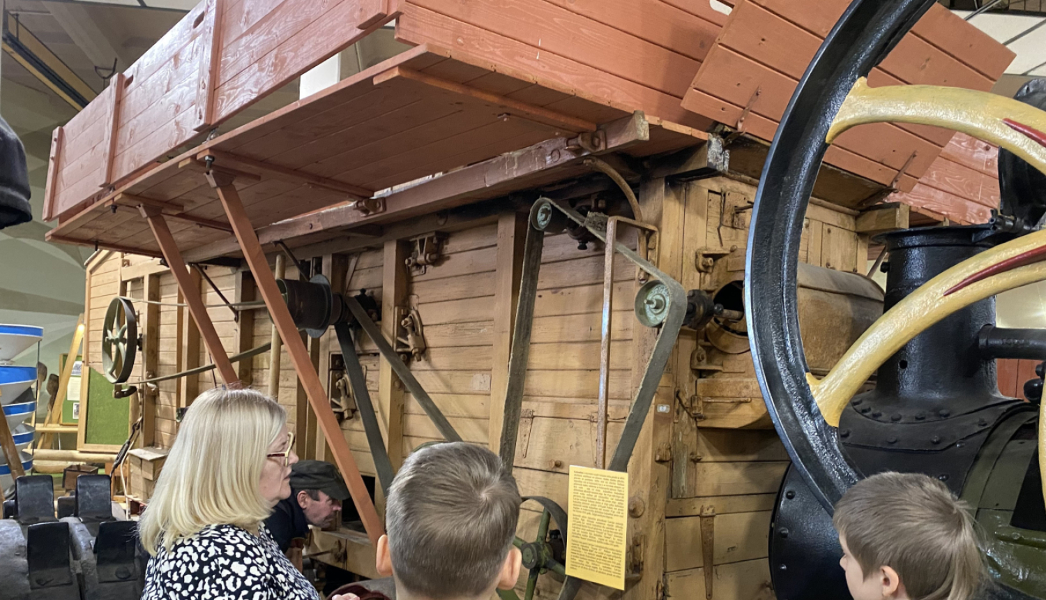 Divi skolnieki muzeja telpās klausās vienā pieaugušajā par senlaicīgu kuļmašīnu