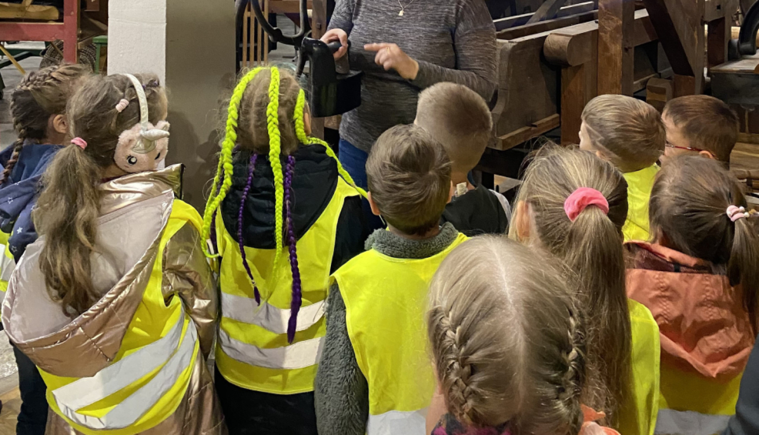 Sieviete gaišiem matiem bērnu bariņā muzeja telpās. Bērni saģērbti zaļās, atstarojošās vestēs