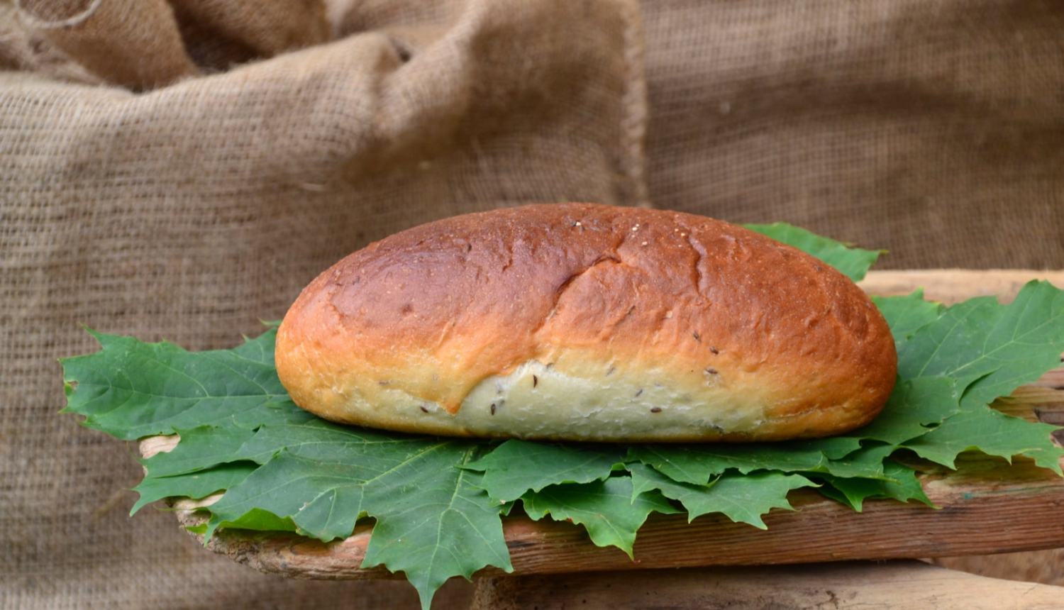 Centrā brūns maizes klaips uz zaļām kļavu lapām, novietots uz maizes lāpstas. Fonā brūni auduma maisi