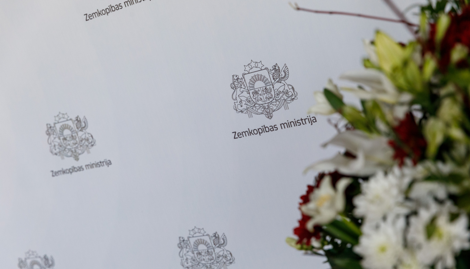 Fonā Zemkopības ministrijas nosaukums atkārtojas uz balta fona. Labajā pusē nedaudz aizmiglots puķu pušķis.