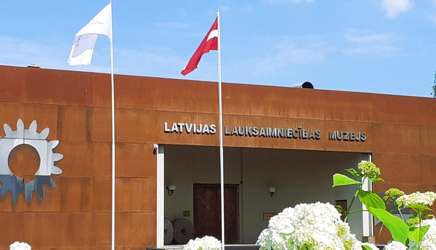  Latvijas Lauksaimniecības muzejs no galvenās ieejas puses. Muzeja priekšā Latvijas un muzeja karogi, kā arī baltas puķes