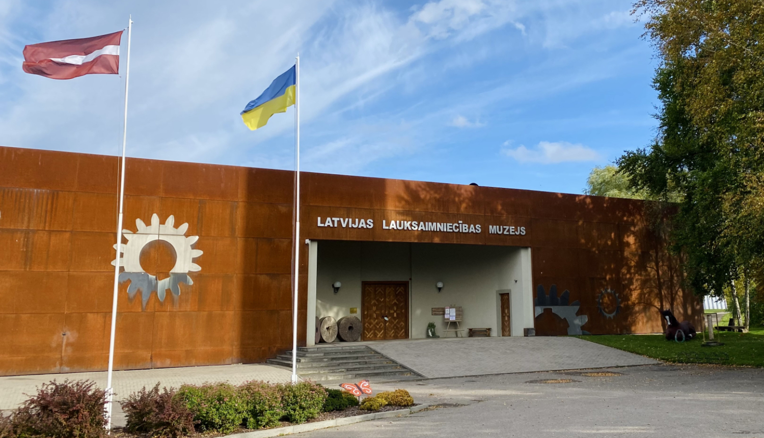 Latvijas Lauksaimniecības muzeja ēkas ieejas skats, ar diviem plīvojošiem karogiem un oranžu taureni pie karogu mastiem